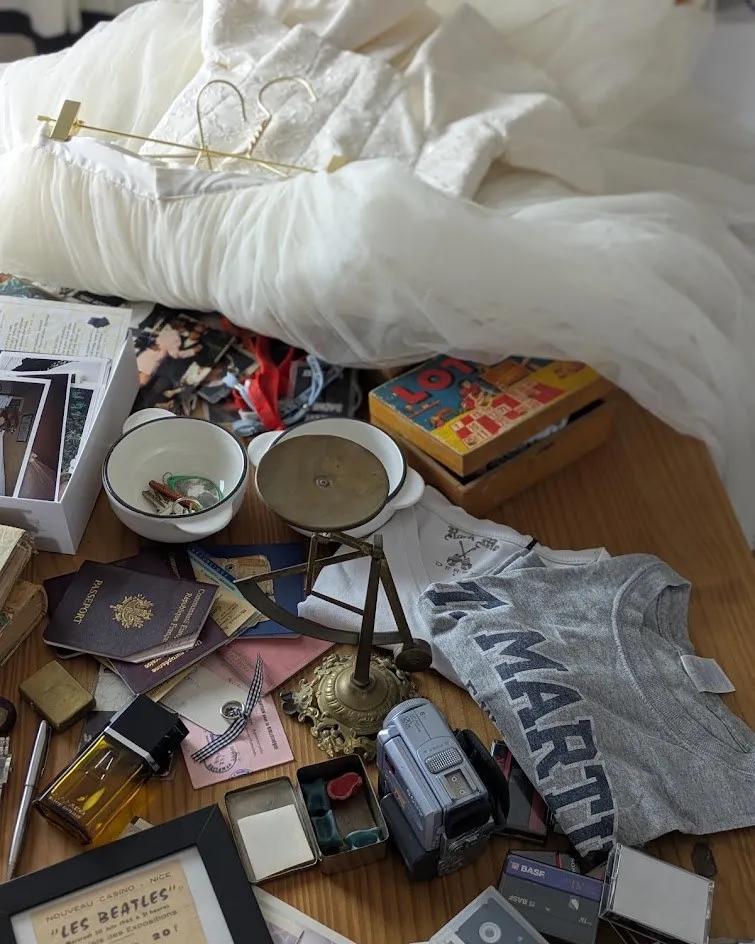 Des souvenirs rassemblés sur un plancher : photos, lunettes, bijoux, cassettes et une robe blanche